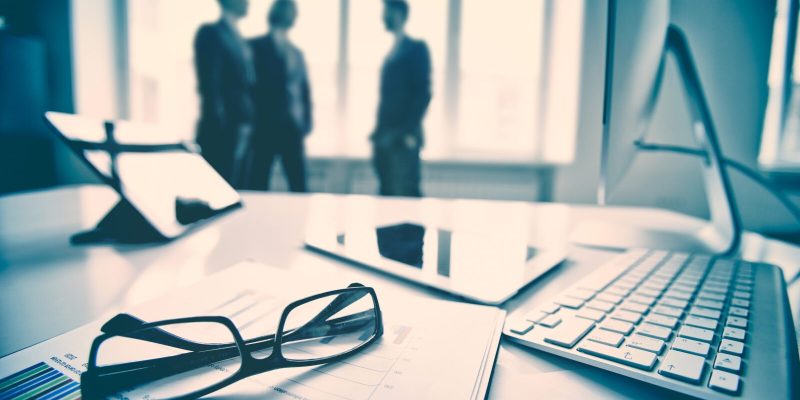 Modern devices, documents, eyeglasses on the foreground, unrecognized businessmen on the background